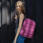 A woman wearing a Backpack Celtic Fucia Pink and Burgundy and Black Highland Plaid, can hold up to a 15" Laptop, or Tablet, many pockets for more, Padded Shoulder Straps leaning against a wooden wall.
