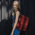 A woman wearing a Celtic Red Highland Plaid backpack leaning against a wooden wall
