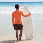 Man in orange shirt holding surfboard on beach.