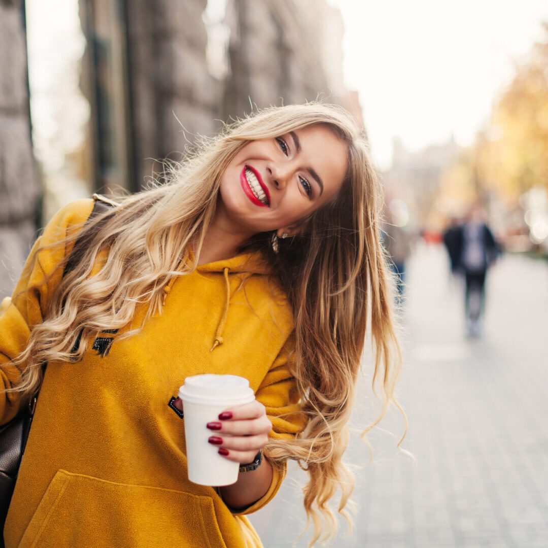 Stylish happy young woman wearing boyfrend jeans, white sneakers bright yellow sweetshot.She holds coffee to go. portrait of smiling girl in sunglasses and bag
